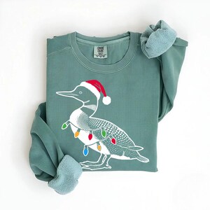 Puede incluir: Una sudadera verde con una ilustraci&oacute;n blanca de un colimbo que lleva un gorro de Pap&aacute; Noel y luces de Navidad. La sudadera tiene cuello redondo y mangas largas.
