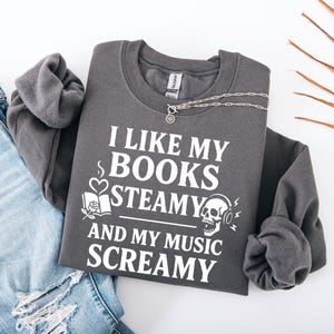May include: Dark gray sweatshirt with the text "I LIKE MY BOOKS STEAMY AND MY MUSIC SCREAMY" in white. The design includes a book, rose, heart, skull with headphones, and musical notes. A silver necklace is draped across the sweatshirt.