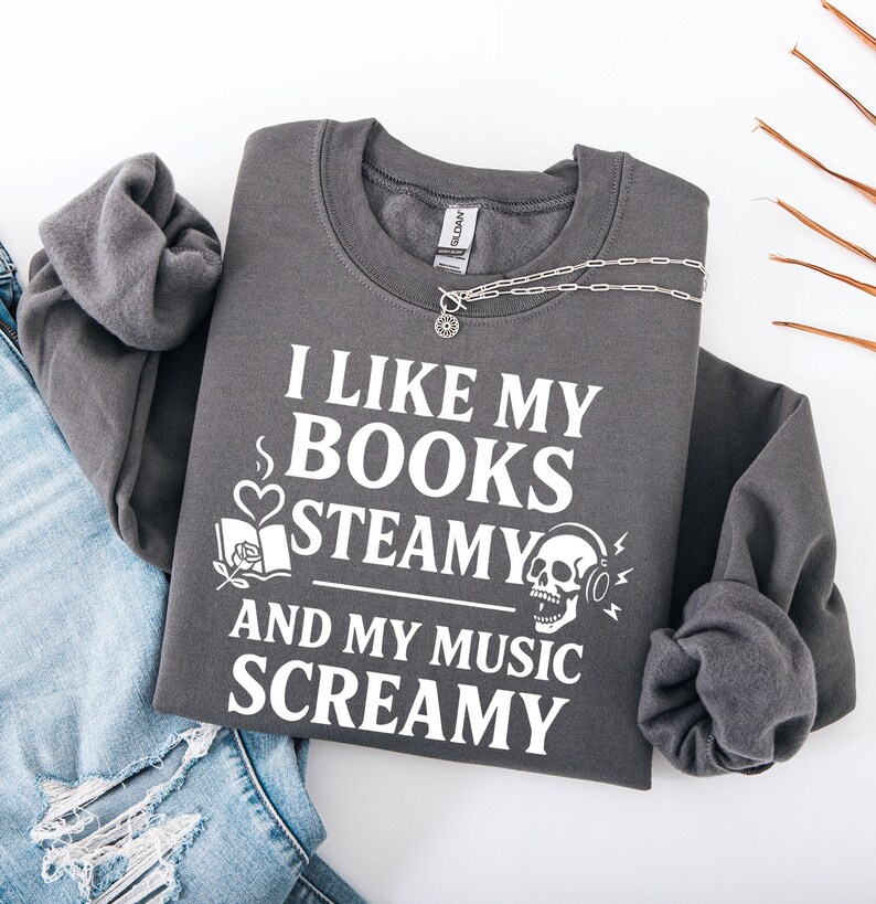 May include: Dark gray sweatshirt with the text "I LIKE MY BOOKS STEAMY AND MY MUSIC SCREAMY" in white. The design includes a book, rose, heart, skull with headphones, and musical notes. A silver necklace is draped across the sweatshirt.