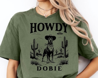 Può includere: T-shirt verde oliva con grafica di un Dobermann con cappello da cowboy e bandana, con la scritta "HOWDY DOBIE". Il design include cactus e un UFO, creando un design unico a tema western.