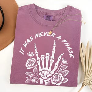 May include: A mauve t-shirt with a white graphic of a skeleton hand making the rock and roll sign with flowers around it. The text "It was never a phase" is written around the hand.