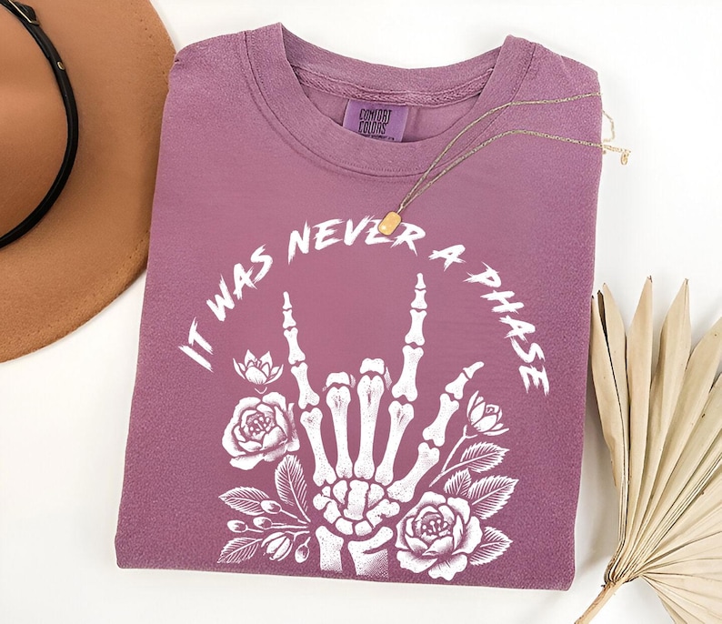 May include: A mauve t-shirt with a white graphic of a skeleton hand making the rock and roll sign with flowers around it. The text "It was never a phase" is written around the hand.