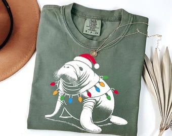 May include: A green t-shirt with a white manatee wearing a Santa hat and Christmas lights. The shirt has the text "COMFORT COLORS" on the tag.