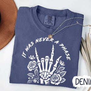 May include: A dark blue t-shirt with a white graphic of a skeleton hand making the rock and roll sign with flowers around it. The text "It Was Never A Phase" is written around the hand. The shirt is labeled "Denim".