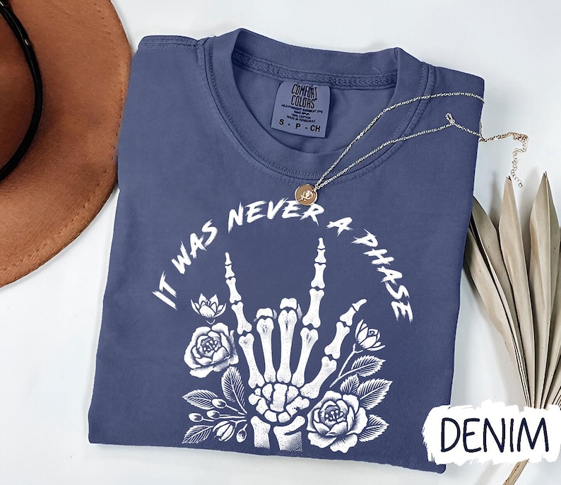 May include: A dark blue t-shirt with a white graphic of a skeleton hand making the rock and roll sign with flowers around it. The text "It Was Never A Phase" is written around the hand. The shirt is labeled "Denim".