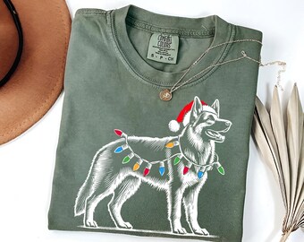 Op de afbeelding: Een groen T-shirt met een witte illustratie van een hond die een kerstmanmuts en kerstverlichting draagt. Het shirt heeft de tekst "COMFORT COLORS" op het label.