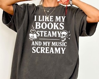 May include: Dark gray t-shirt with the text "I LIKE MY BOOKS STEAMY AND MY MUSIC SCREAMY" in white. The design includes a book, heart, and skull with headphones. The shirt has short sleeves and a relaxed fit.