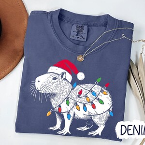 May include: A navy blue t-shirt with a white illustration of a capybara wearing a Santa hat and Christmas lights. The text "DENIM" is visible in the bottom right corner of the image.