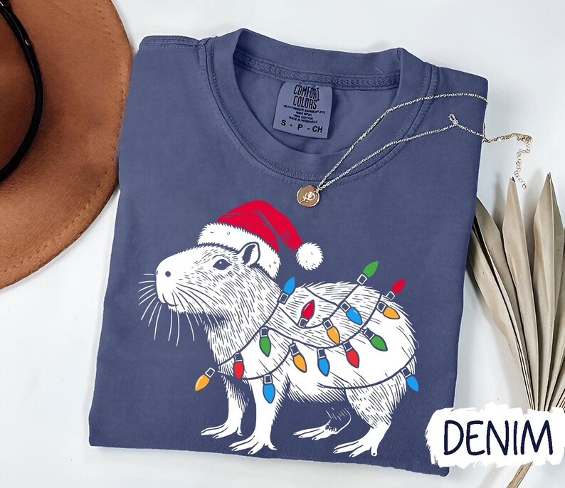 May include: A navy blue t-shirt with a white illustration of a capybara wearing a Santa hat and Christmas lights. The text "DENIM" is visible in the bottom right corner of the image.