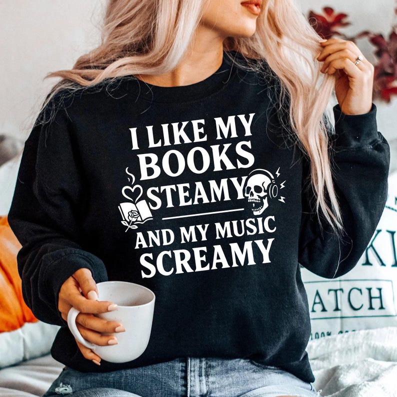May include: Black sweatshirt with the text "I LIKE MY BOOKS STEAMY AND MY MUSIC SCREAMY" in white. The design includes a book, heart, skull, and headphones. A person is holding a white mug.