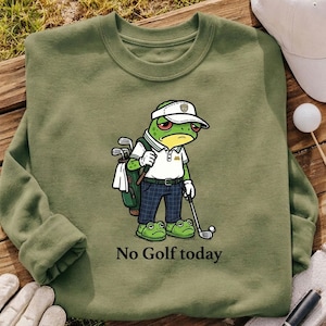 May include: Olive green sweatshirt featuring a cartoon frog golfer with a visor, golf bag, and club. The frog is wearing a white polo shirt and plaid pants. Text on the sweatshirt reads "No Golf today". A golf ball and hat are also visible.
