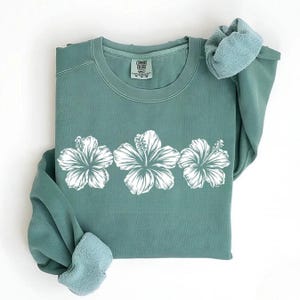May include: A teal sweatshirt with three white hibiscus flower designs. The sweatshirt has a crew neck and long sleeves with a soft, fleece-like lining. The tag reads "Comfort Colors".