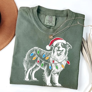 Puede incluir: Una camiseta verde oliva con un gráfico de un perro con un gorro de Papá Noel y luces navideñas. El perro es blanco con detalles negros. También se ve un collar plateado y un sombrero marrón.