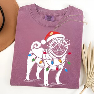 May include: A purple t-shirt with a white pug wearing a Santa hat and Christmas lights. The shirt has the text "Comfort Colors" on the tag.