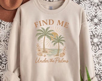 Może przedstawiać: Beżowa bluza z tropikalną grafiką. Projekt przedstawia dwie palmy, słońce i napis "FIND ME" nad "Under the Palms". Grafika jest w odcieniach brązu i zieleni.