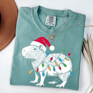 May include: A green t-shirt with a white illustration of a capybara wearing a Santa hat and Christmas lights. The text "COMFORT COLORS" is printed on the tag.