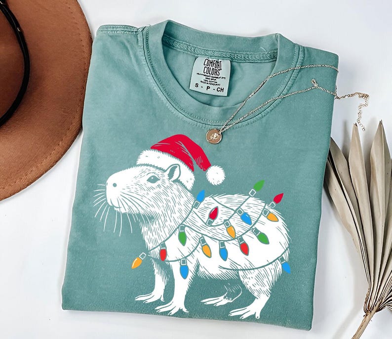 May include: A green t-shirt with a white illustration of a capybara wearing a Santa hat and Christmas lights. The text "COMFORT COLORS" is printed on the tag.