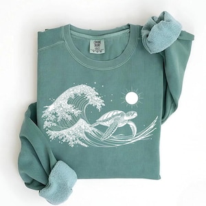 May include: A teal green sweatshirt with a white graphic of a sea turtle swimming through a wave. The graphic also includes a sun and stars. The sweatshirt has the text "COMMON COLORS" on the tag.