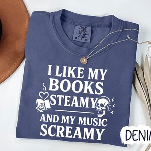 May include: A denim blue t-shirt with the text "I LIKE MY BOOKS STEAMY AND MY MUSIC SCREAMY" in white. The graphic includes a book, a skull with headphones, and a heart with steam. A gold necklace is draped across the shirt.