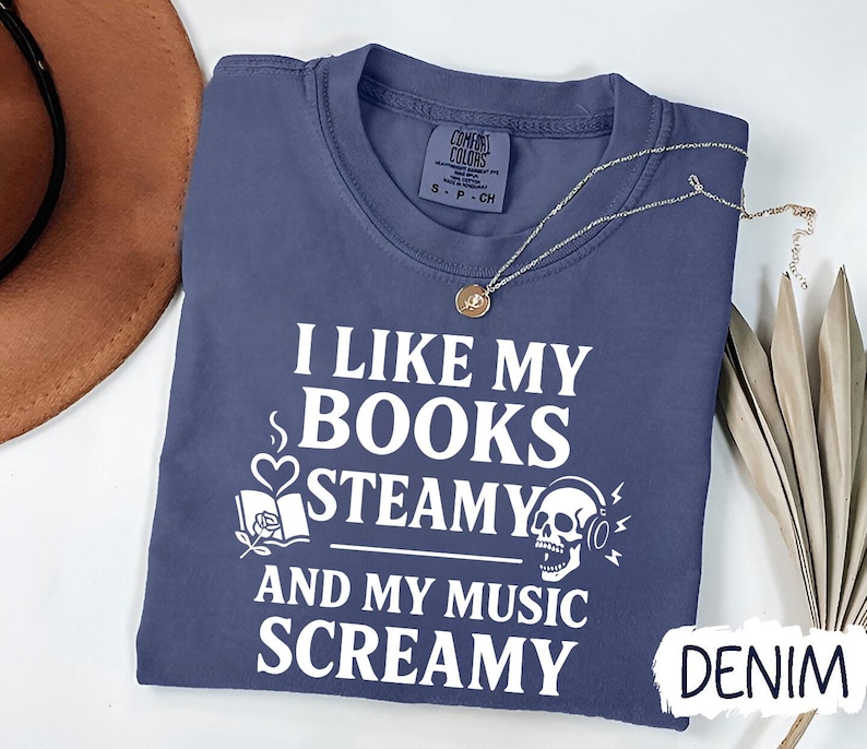 May include: A denim blue t-shirt with the text "I LIKE MY BOOKS STEAMY AND MY MUSIC SCREAMY" in white. The graphic includes a book, a skull with headphones, and a heart with steam. A gold necklace is draped across the shirt.