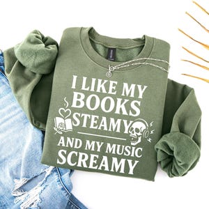 May include: An olive green sweatshirt with the text "I LIKE MY BOOKS STEAMY AND MY MUSIC SCREAMY" in white. The design includes a heart, a book, and a skull with headphones. A silver necklace is draped across the sweatshirt.