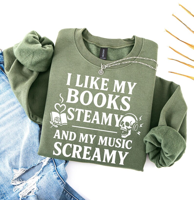 May include: An olive green sweatshirt with the text "I LIKE MY BOOKS STEAMY AND MY MUSIC SCREAMY" in white. The design includes a heart, a book, and a skull with headphones. A silver necklace is draped across the sweatshirt.
