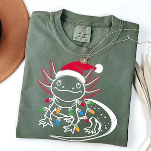 May include: A green t-shirt featuring a white axolotl wearing a Santa hat and Christmas lights. The tag reads "COMFORT COLORS".