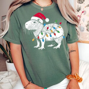 May include: A green short-sleeved t-shirt with a white illustration of a capybara wearing a Santa hat and Christmas lights.
