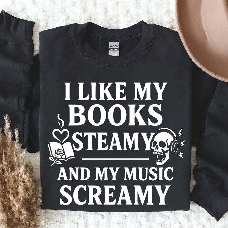 May include: Black sweatshirt with white text: "I LIKE MY BOOKS STEAMY AND MY MUSIC SCREAMY." Graphic includes a book, heart, and skull with headphones. A cozy apparel item for book and music lovers.