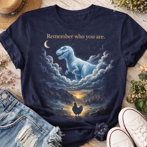 Puede incluir: Camiseta azul marino con un diseño de Tyrannosaurus Rex entre nubes, una luna creciente y el texto "Remember who you are." Un pollo está en primer plano. La camiseta se combina con pantalones cortos de mezclilla y zapatillas blancas.