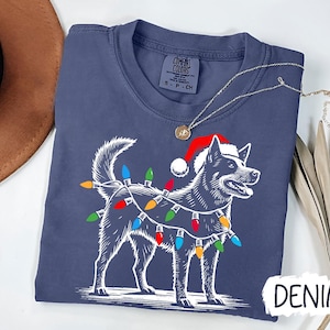 Puede incluir: Una camiseta azul oscuro con una ilustración blanca de un perro con un gorro de Papá Noel y luces de Navidad. El texto "DENIM" está impreso en blanco en la esquina inferior derecha de la camiseta.