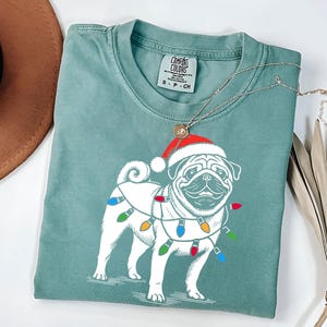 May include: A teal blue t-shirt with a white pug wearing a red Santa hat and Christmas lights around its neck. The shirt has a tag that says "COMFORT COLORS" and "S - P - CH".