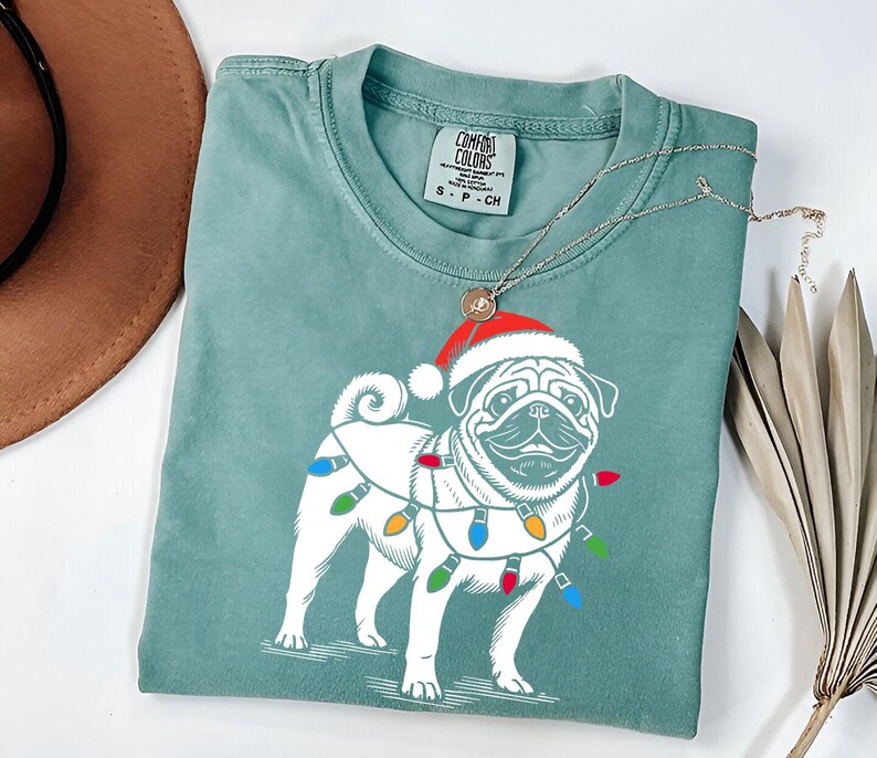 May include: A teal blue t-shirt with a white pug wearing a red Santa hat and Christmas lights around its neck. The shirt has a tag that says "COMFORT COLORS" and "S - P - CH".