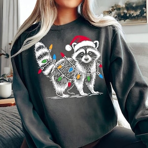 Puede incluir: Una sudadera gris oscuro con un gr&aacute;fico blanco de un mapache con un gorro de Pap&aacute; Noel y rodeado de luces navide&ntilde;as de colores.