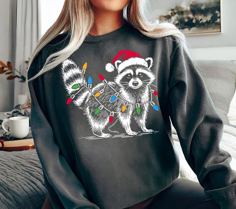 Puede incluir: Una sudadera gris oscuro con un gr&aacute;fico blanco de un mapache con un gorro de Pap&aacute; Noel y rodeado de luces navide&ntilde;as de colores.