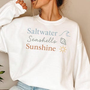 以下が含まれることがあります： 白のスウェットシャツ。青と茶色の文字で「Saltwater, Seashells, Sunshine」と書かれています。波、貝殻、太陽のグラフィックが含まれています。