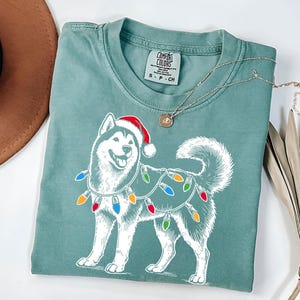 Puede incluir: Una camiseta azul turquesa con una ilustración blanca de un perro husky con un gorro de Papá Noel y luces de Navidad. La camiseta tiene el texto "COMFORT COLORS" en la etiqueta.