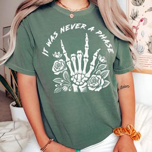 May include: Green t-shirt with a white graphic of a skeleton hand making the rock and roll sign with flowers around it. The text on the shirt reads "It was never a phase."