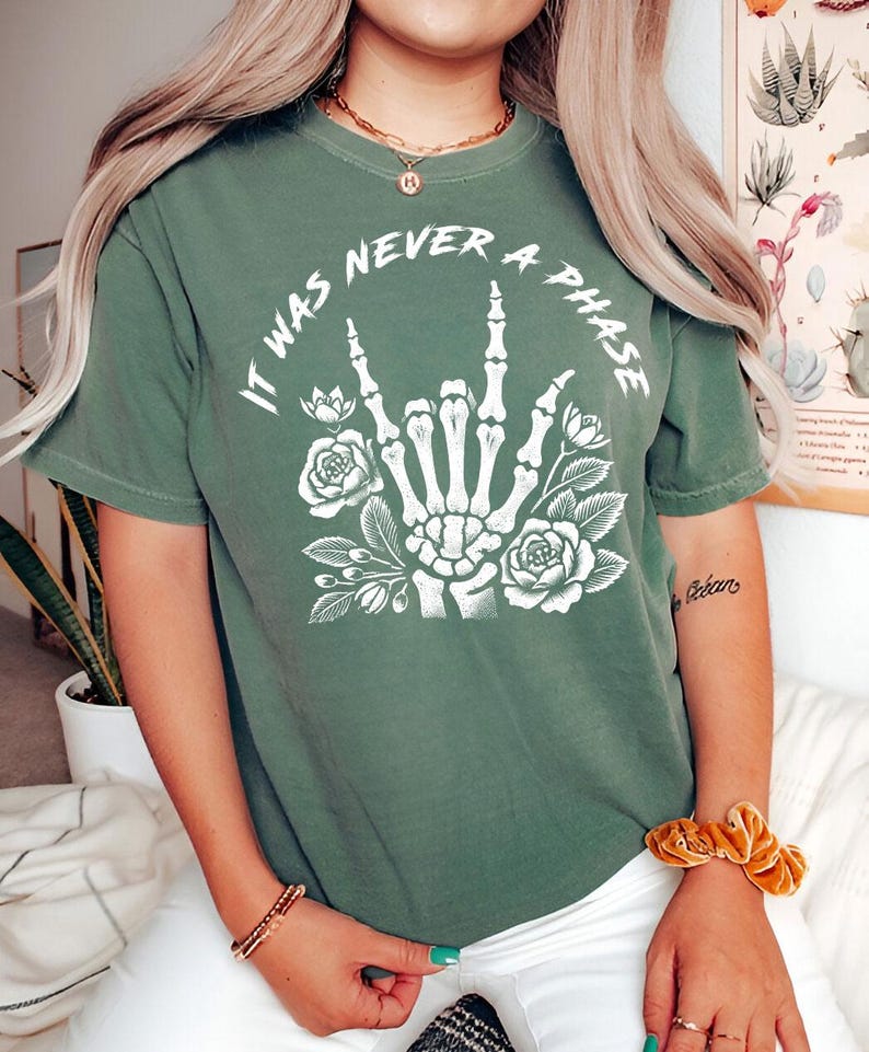 May include: Green t-shirt with a white graphic of a skeleton hand making the rock and roll sign with flowers around it. The text on the shirt reads "It was never a phase."