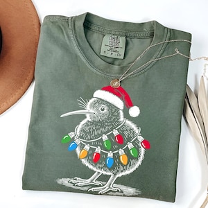 Puede incluir: Una camiseta verde con una ilustración blanca de un kiwi con un gorro de Papá Noel y luces de Navidad. La camiseta tiene el texto "COMFORT COLORS" en la etiqueta.