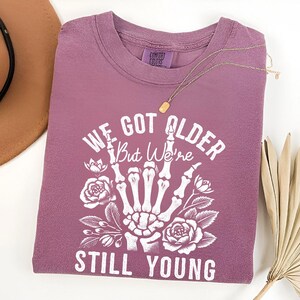 Pode incluir: Uma camiseta malva com um gr&aacute;fico branco de uma m&atilde;o de esqueleto mostrando o dedo do meio. O gr&aacute;fico tamb&eacute;m inclui flores e o texto "We Got Older But We're Still Young".