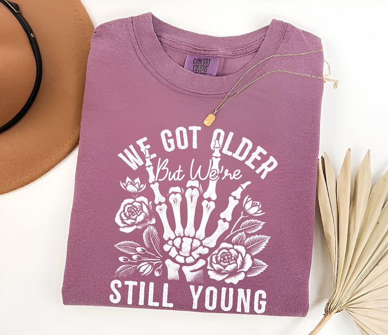 Pode incluir: Uma camiseta malva com um gr&aacute;fico branco de uma m&atilde;o de esqueleto mostrando o dedo do meio. O gr&aacute;fico tamb&eacute;m inclui flores e o texto "We Got Older But We're Still Young".