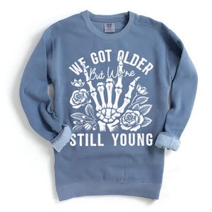 May include: A blue sweatshirt with a white graphic print of a skeleton hand with flowers and the text "We Got Older But We're Still Young".