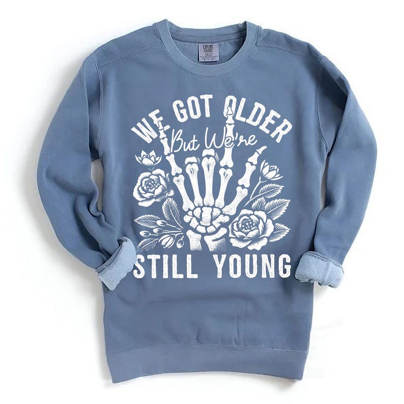 May include: A blue sweatshirt with a white graphic print of a skeleton hand with flowers and the text "We Got Older But We're Still Young".