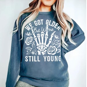 May include: A blue sweatshirt with a white graphic print of a skeleton hand giving the middle finger with flowers and the text "We got older but we're still young".