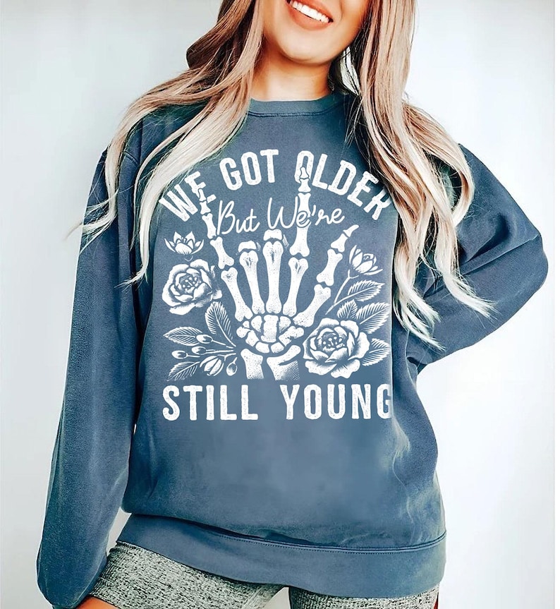 May include: A blue sweatshirt with a white graphic print of a skeleton hand giving the middle finger with flowers and the text "We got older but we're still young".