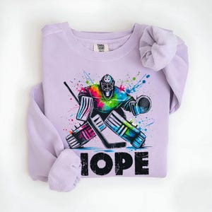 May include: Purple sweatshirt with a colorful graphic of a hockey goalie in action. The text "HOPE" is printed below the graphic.