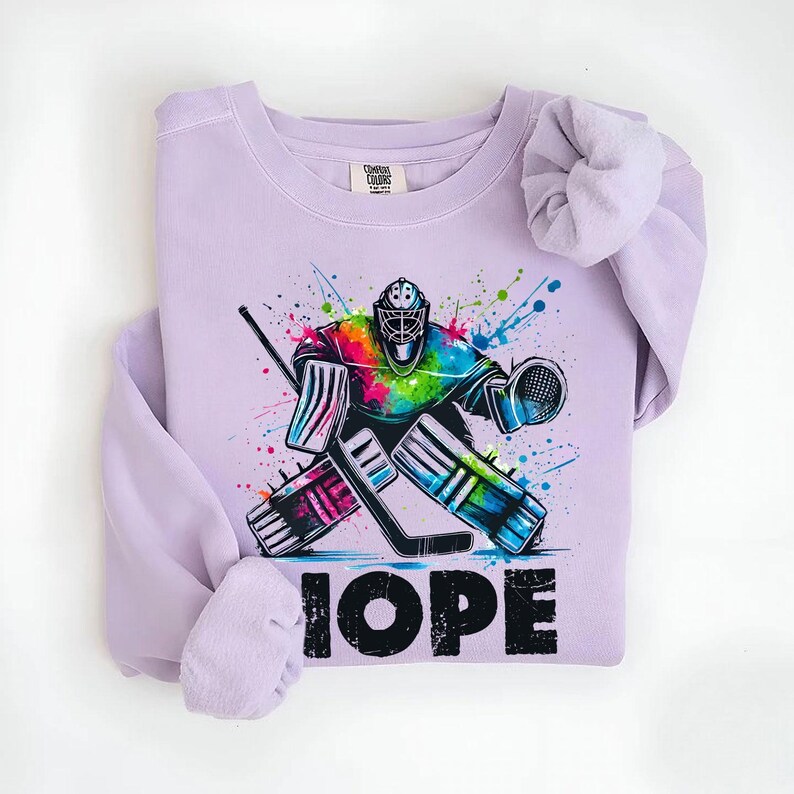 May include: Purple sweatshirt with a colorful graphic of a hockey goalie in action. The text "HOPE" is printed below the graphic.