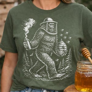 Könnte beinhalten: Olivgrünes T-Shirt mit weißer Grafik eines Bigfoot-Imkers. Der Bigfoot trägt einen Imkerhut und hält einen Raucher und einen Bienenstock. Bienen fliegen um den Bigfoot herum. Ein Glas Honig und ein Honiglöffel sind im Hintergrund.