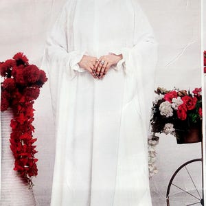 May include: A white abaya with long sleeves and a long skirt. The abaya is made of a soft, flowing fabric. The woman is wearing a white headscarf.